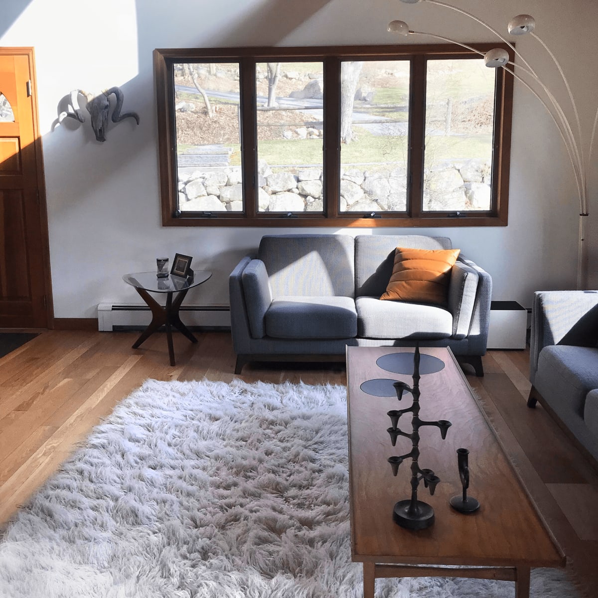 A bright living room featuring two gray sofas, a wooden coffee table, a white rug, and large windows that overlook a stone wall and outdoor space.