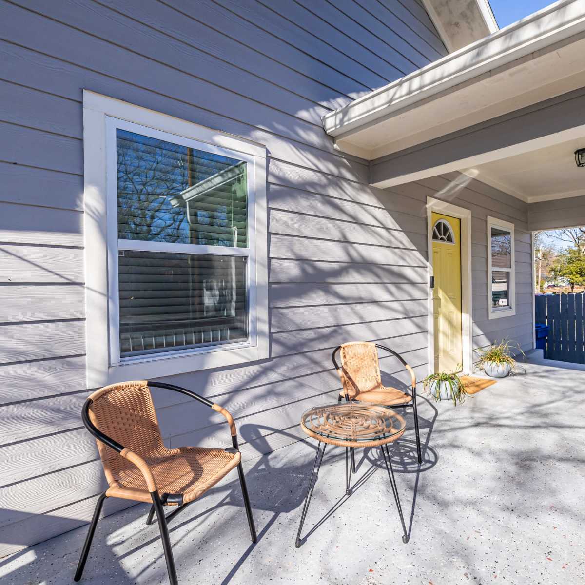 A porch with two wicker chairs and a small round table, alongside a yellow front door and a gray house exterior.