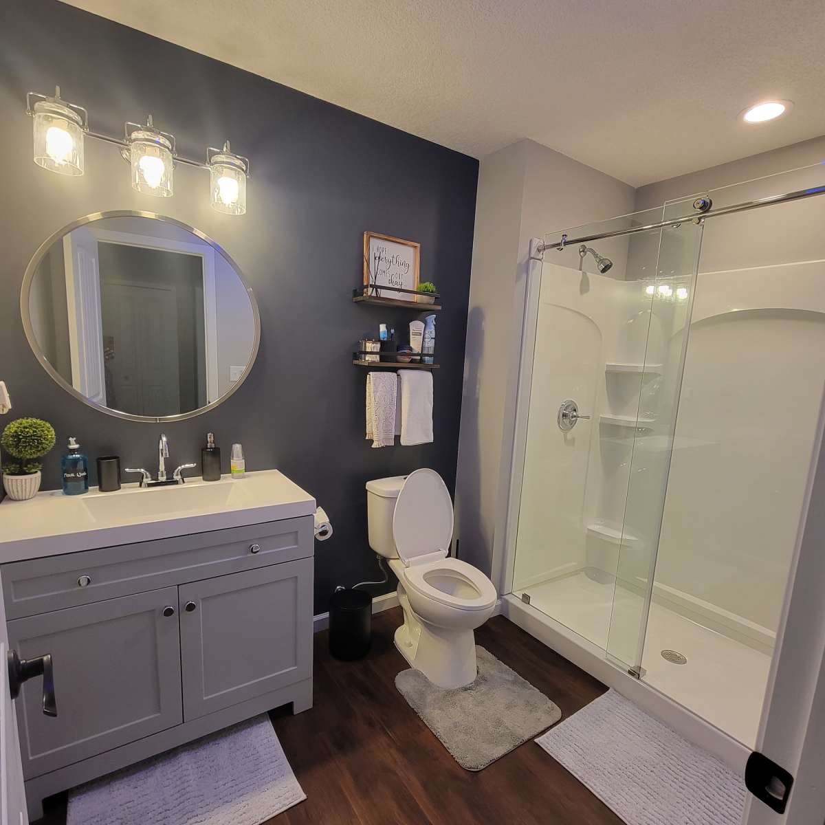 The bathroom features a modern design with a circular mirror, a gray vanity, a glass shower, and wood-like flooring.