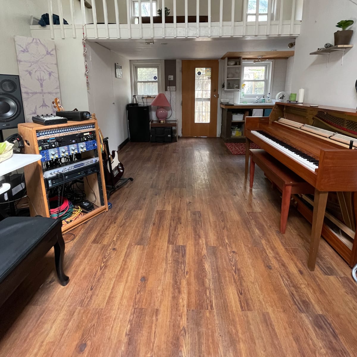 A spacious room with wooden flooring, including a piano, a sound equipment rack, and a loft area above.
