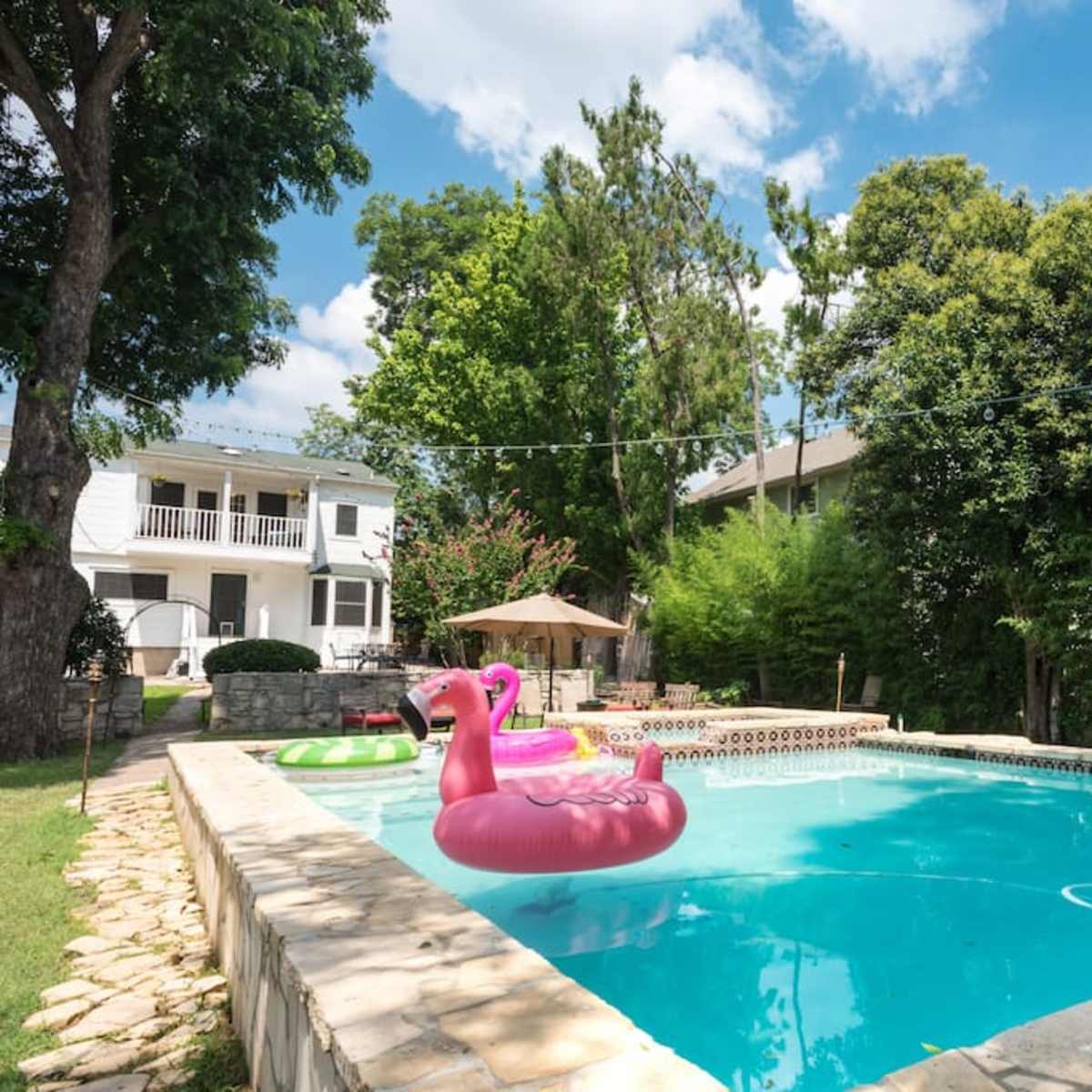 A swimming pool with pink flamingo floaties surrounded by greenery and a two-story house in the background.
