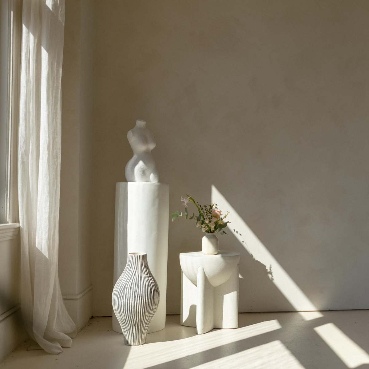 A minimalist indoor space with three white decorative pieces, including a sculpture and vases, illuminated by natural light filtering through a window.