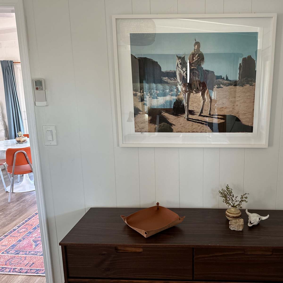 A framed photograph of a person on horseback in a desert landscape is hung on a white-paneled wall above a wooden dresser with decorative items.