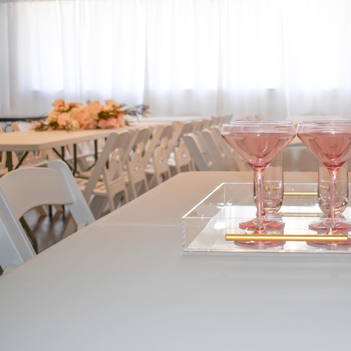 A clear tray holds two pink glasses on a table surrounded by white chairs in a brightly lit room.