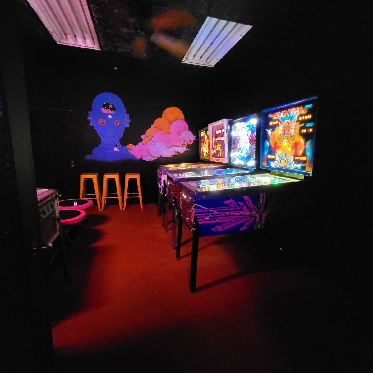 A dimly lit room features several pinball machines and colorful stools against a black wall with a mural.