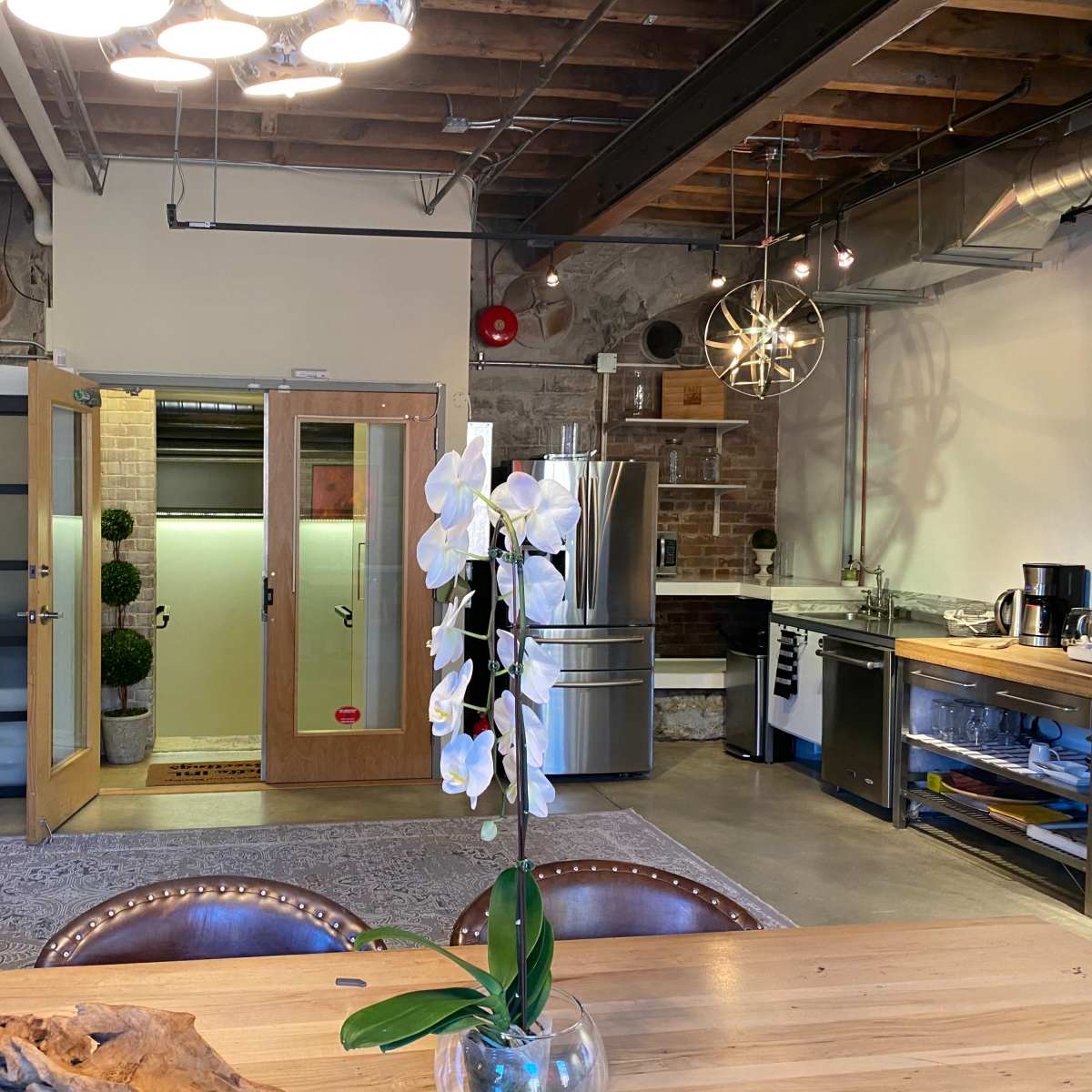 A modern kitchen and dining area with a wooden table, flowering plant, metal appliances, and a door leading to a separate room.