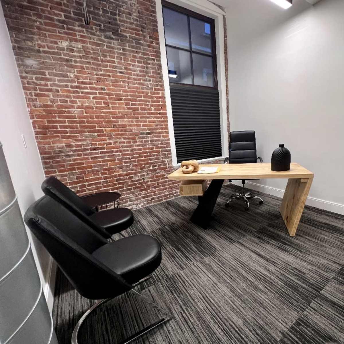 A small office with a wooden desk, a black chair, and two black lounge chairs against a brick wall.