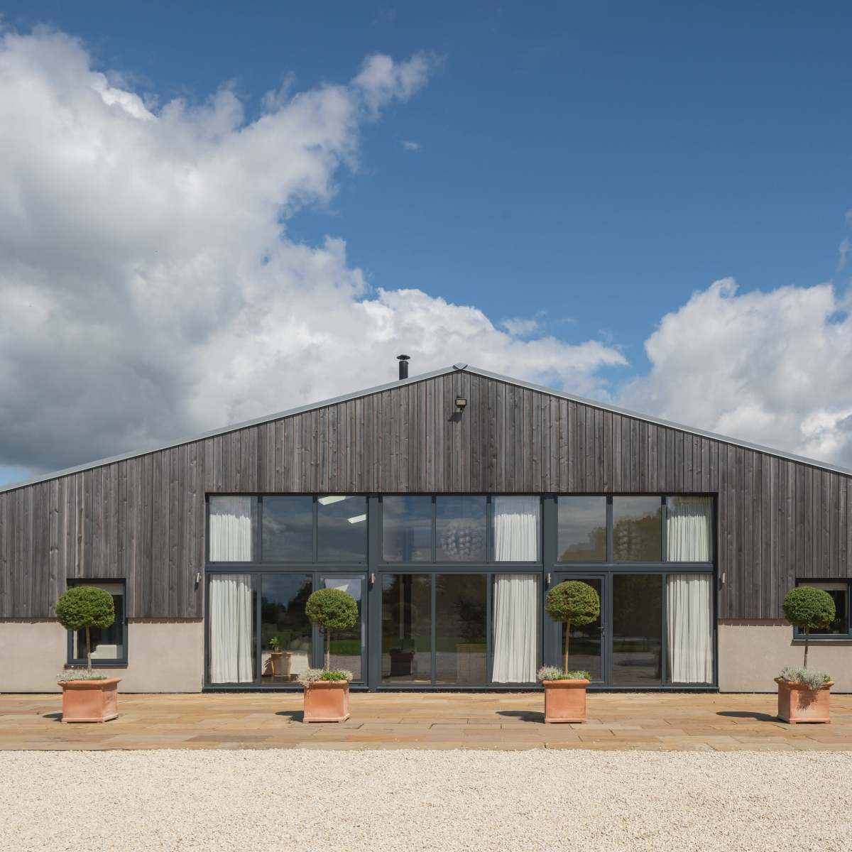 A modern barn-style house with a wooden facade, large glass windows, and potted topiary plants in front, set against a cloudy sky.