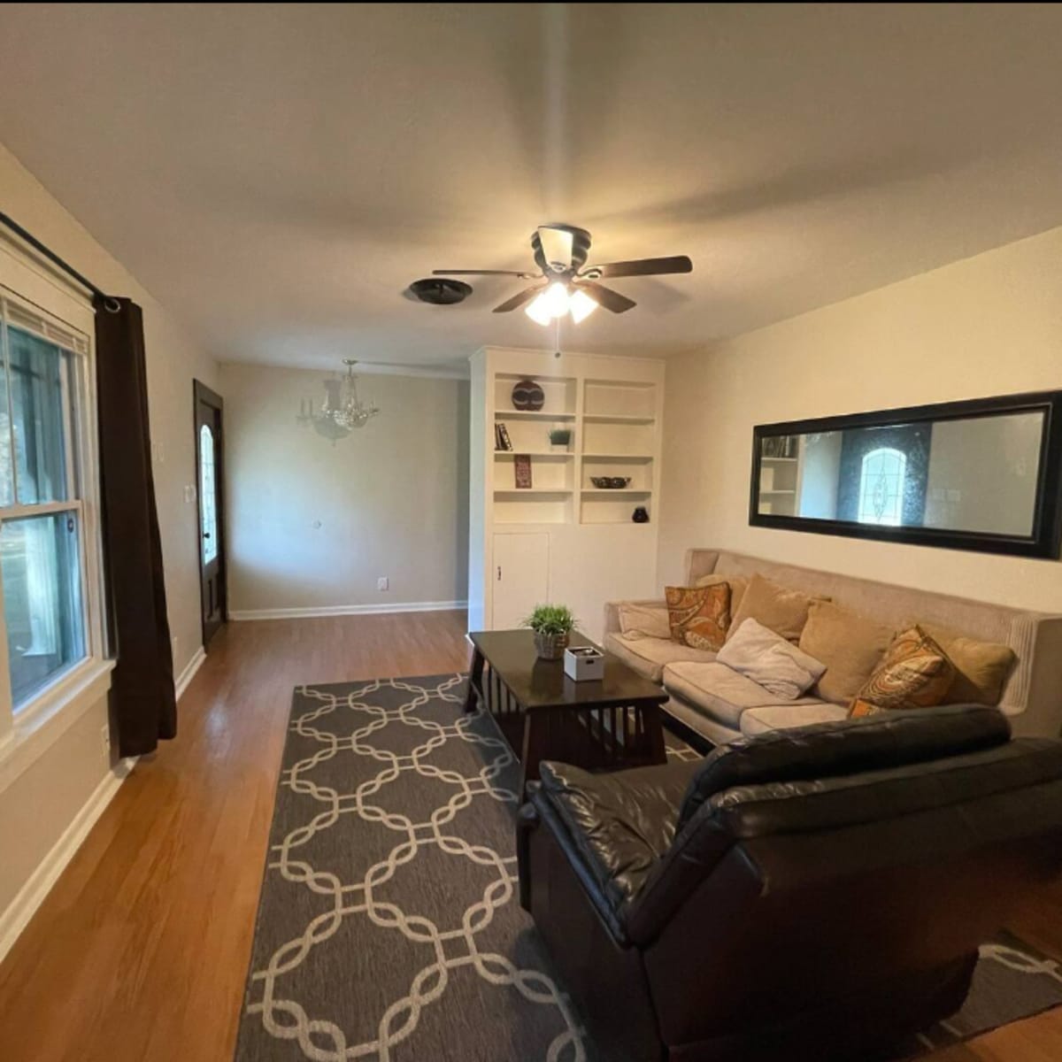 A living room with a beige sofa, a black recliner, a coffee table, and large windows allowing natural light, along with shelves and a chandelier.