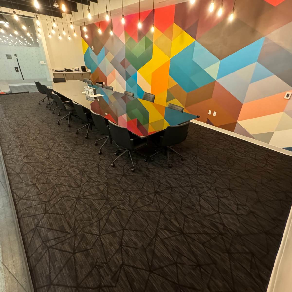 A conference room features a large glass table surrounded by black chairs, with a colorful geometric mural on the wall and modern lighting fixtures above.