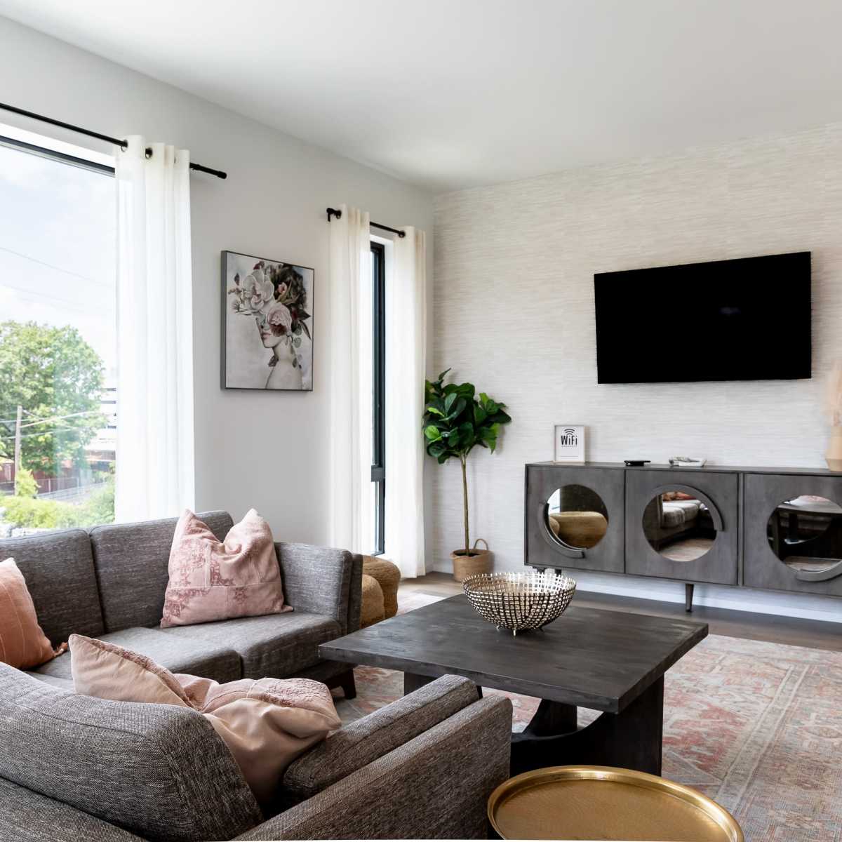 A modern living room features a gray sectional sofa, a wooden coffee table, and a wall-mounted television, with large windows allowing natural light to fill the space.