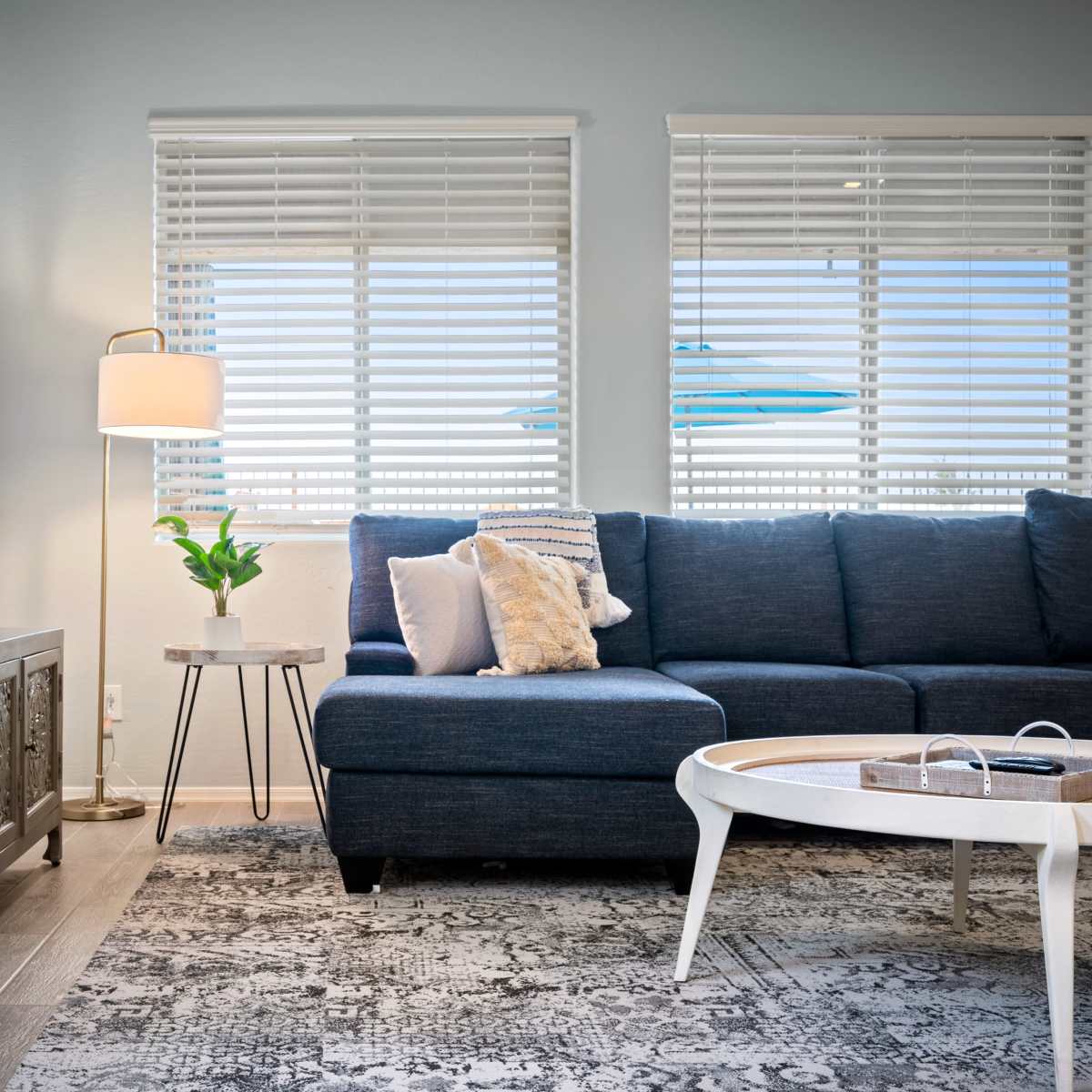 A modern living room with a blue sectional sofa, a coffee table, a television mounted on the wall, and two windows with blinds letting in natural light.