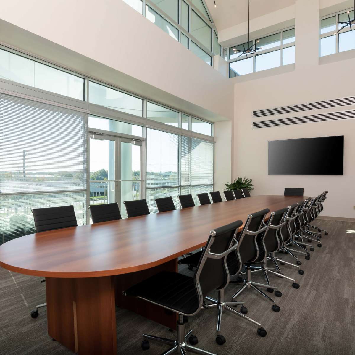 A spacious conference room featuring a long wooden table surrounded by black chairs, large windows with blinds, and a wall-mounted screen.