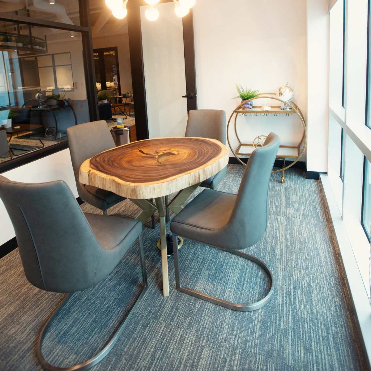 A modern office space features a round wooden table with four grey chairs and a stylish light fixture above.