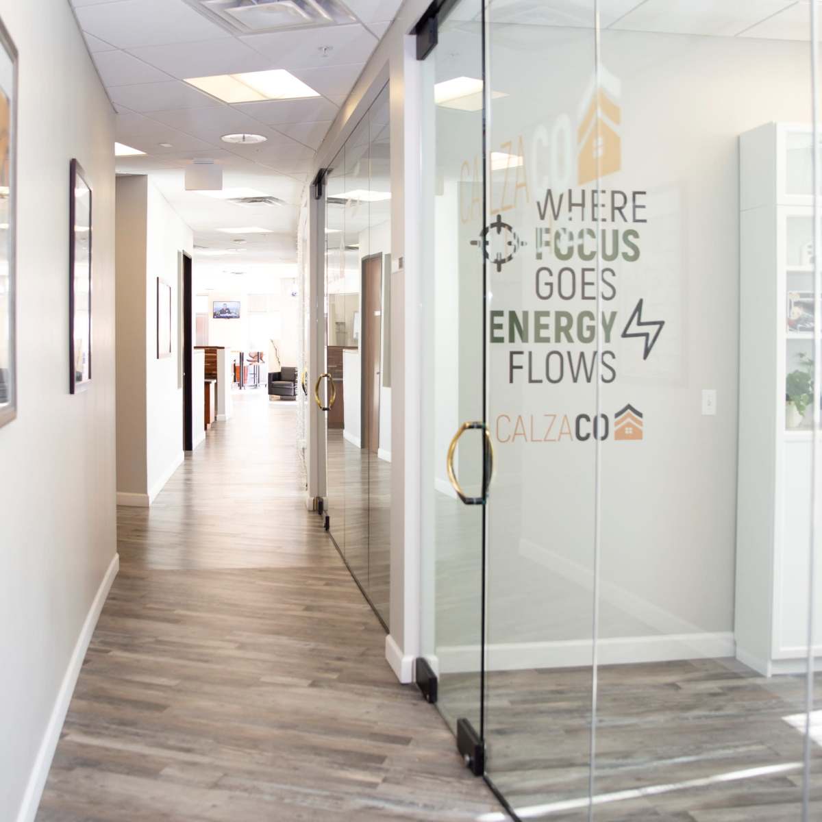 A long office corridor features glass doors with a motivational slogan and wooden flooring leading to various workspaces.