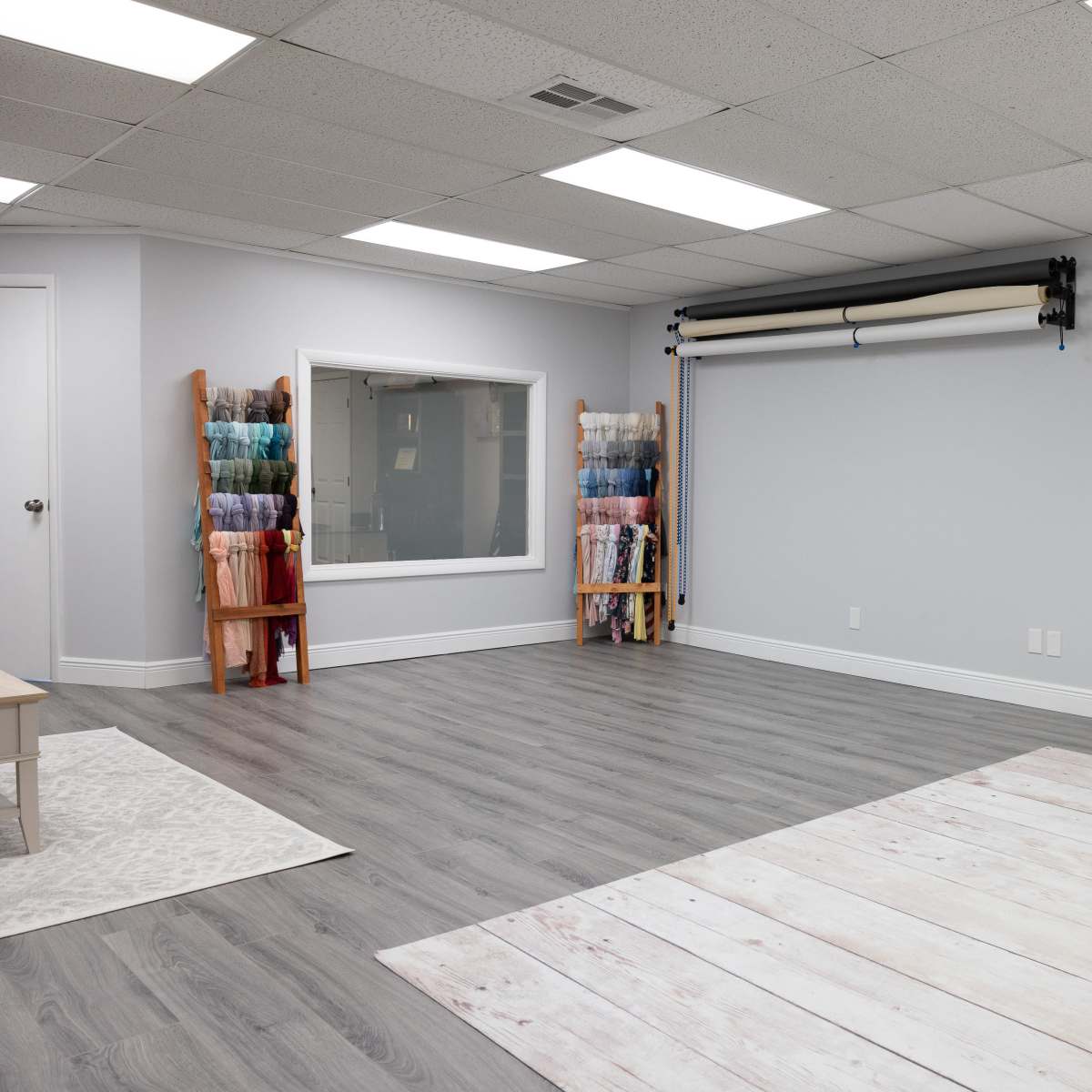 A spacious studio with gray walls, a large window, and shelves displaying rolls of fabric along with a door and a couch in the corner.