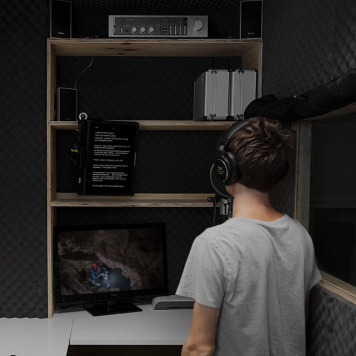 A person wearing headphones stands in a soundproof recording booth, facing a monitor and a shelf with audio equipment.