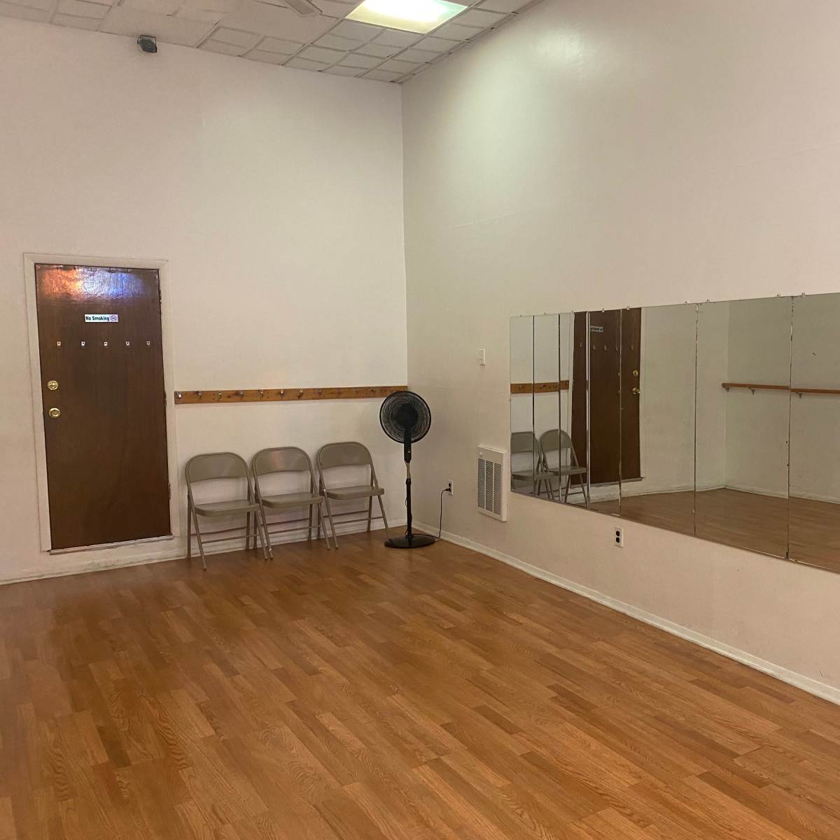 A dance studio with a wooden floor, large mirrors on one wall, and a row of folding chairs beside a door.