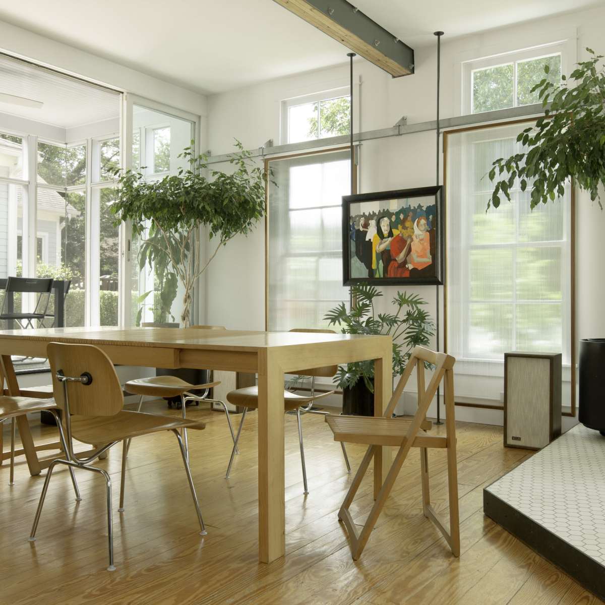A well-lit dining area featuring a wooden table surrounded by chairs, with large windows, potted plants, and a painting on the wall.