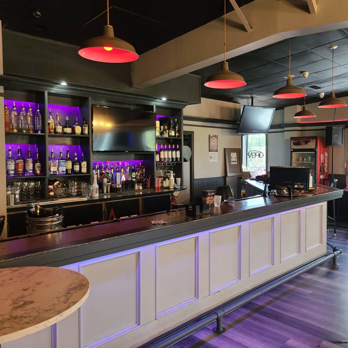 A modern bar area with a long countertop, a selection of spirits on shelves, and hanging red lights illuminating the space.