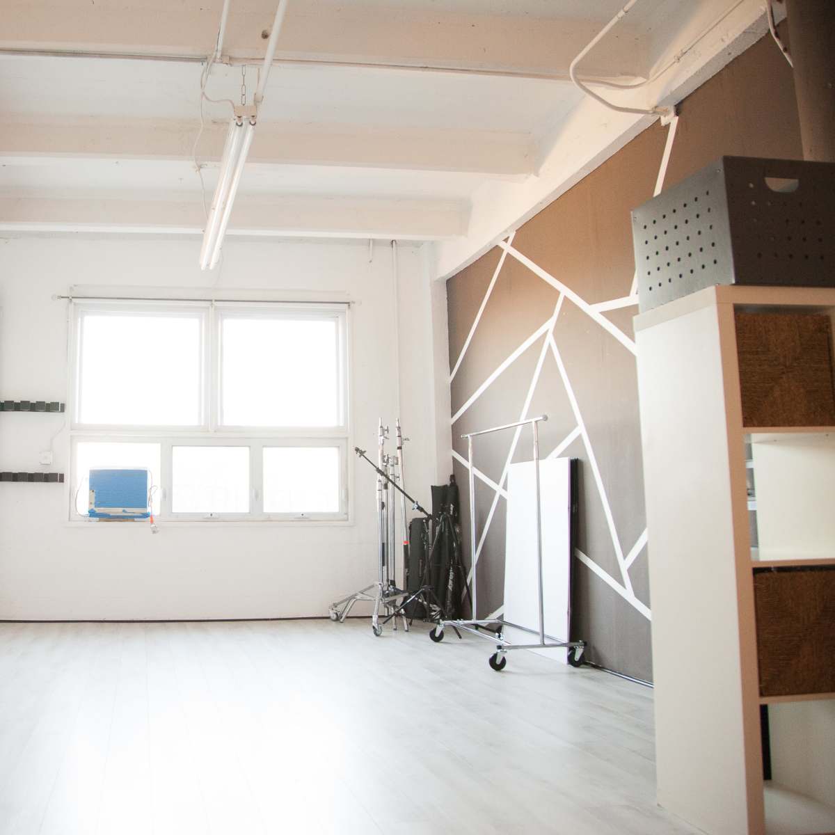 A spacious, well-lit studio with a white floor, a large window, and various equipment including a backdrop stand and storage units.