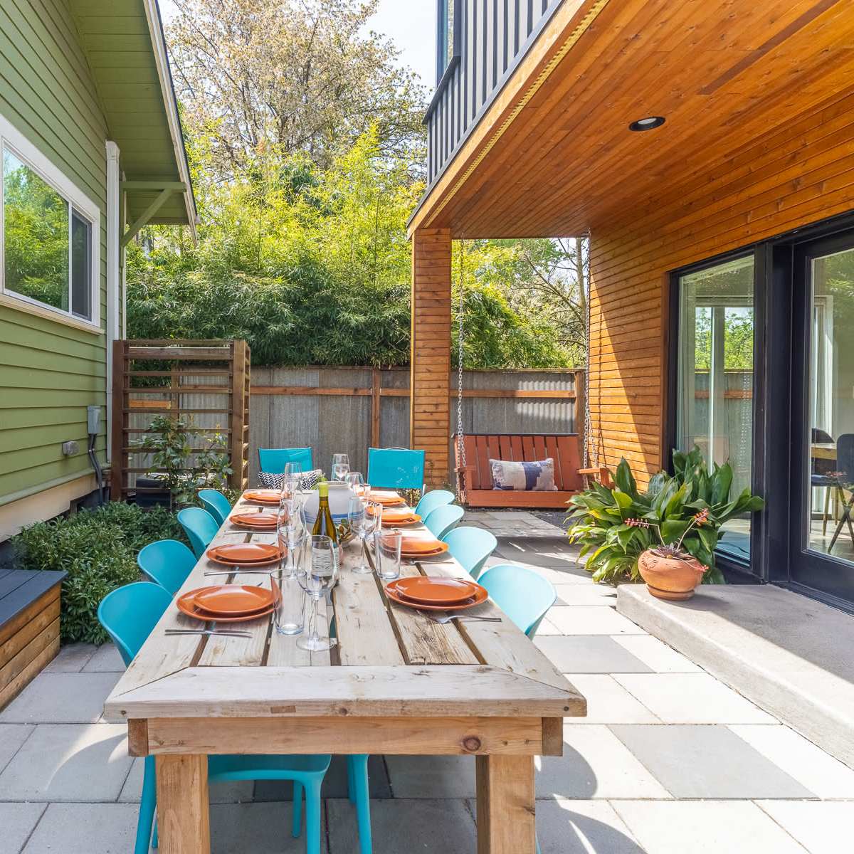 A wooden dining table set with plates and glasses is arranged in an outdoor patio between a green house and a modern wooden structure.