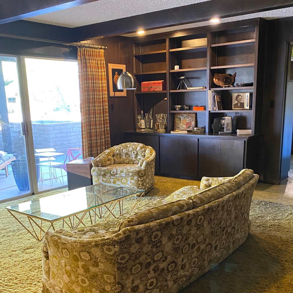 A living room with a patterned sofa and two armchairs facing a coffee table, with a bookshelf and large glass doors leading to an outdoor space.