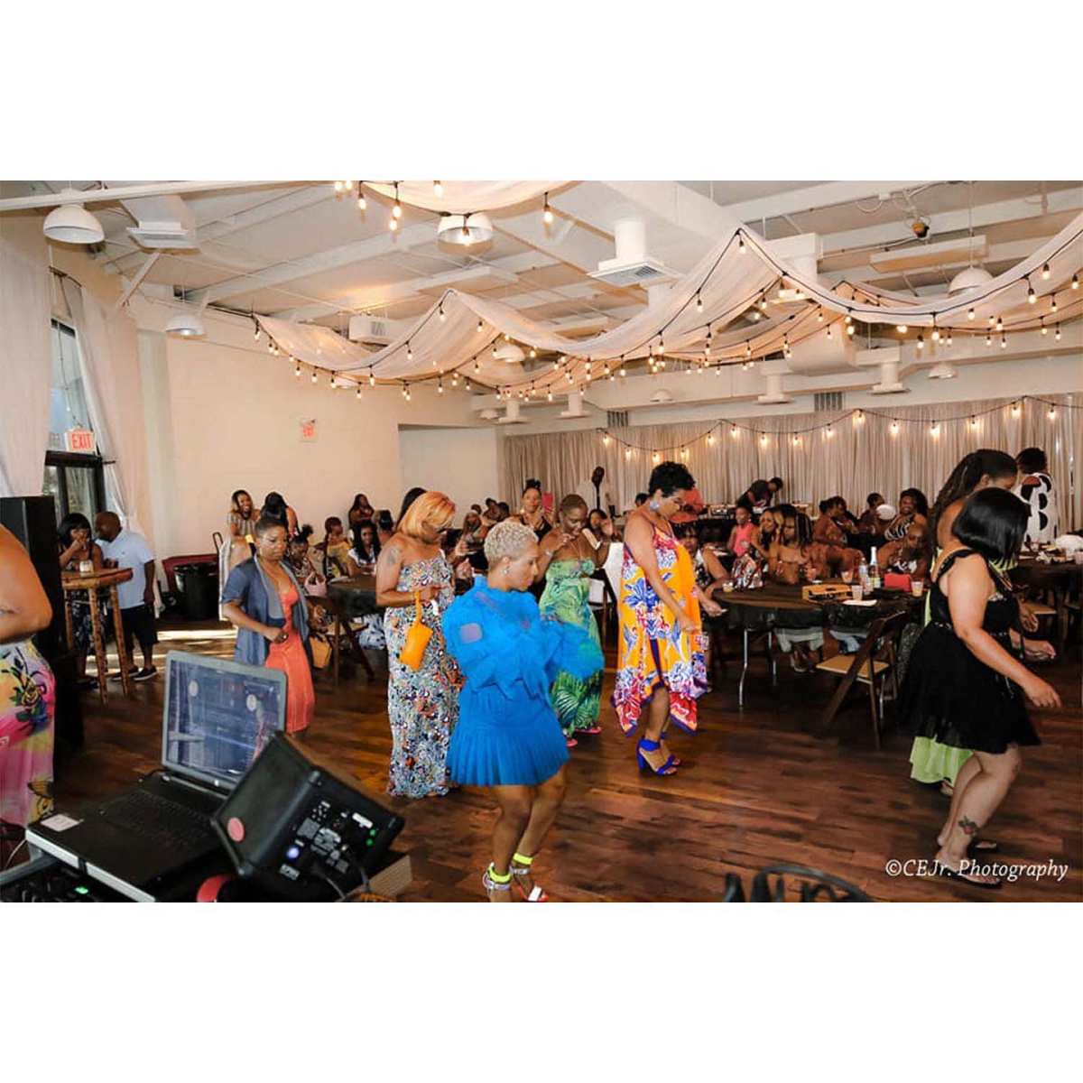 A group of people dances in a decorated venue filled with tables and string lights.