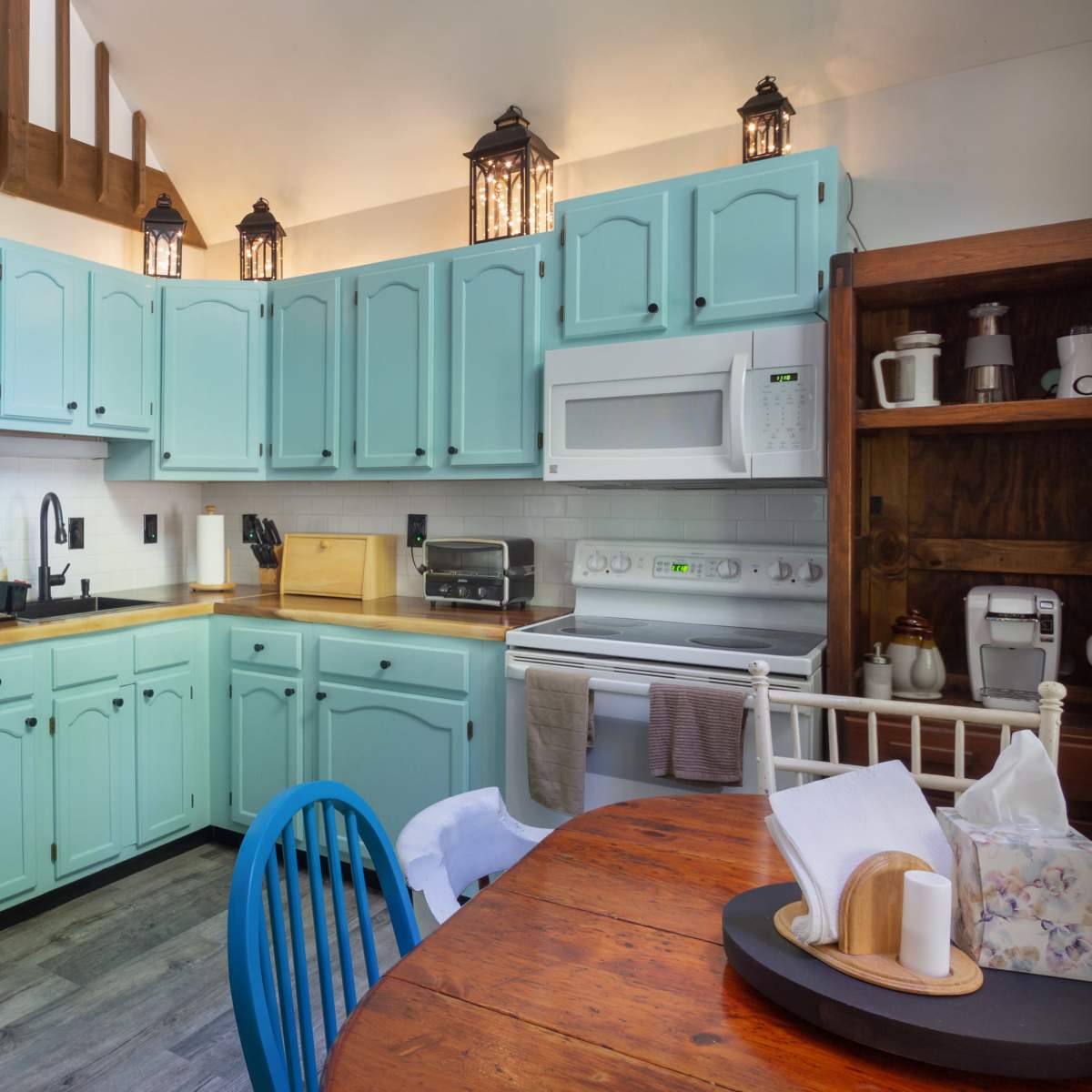 A kitchen with turquoise cabinets, a white refrigerator, a microwave, a stove, and a dining table with blue chairs.