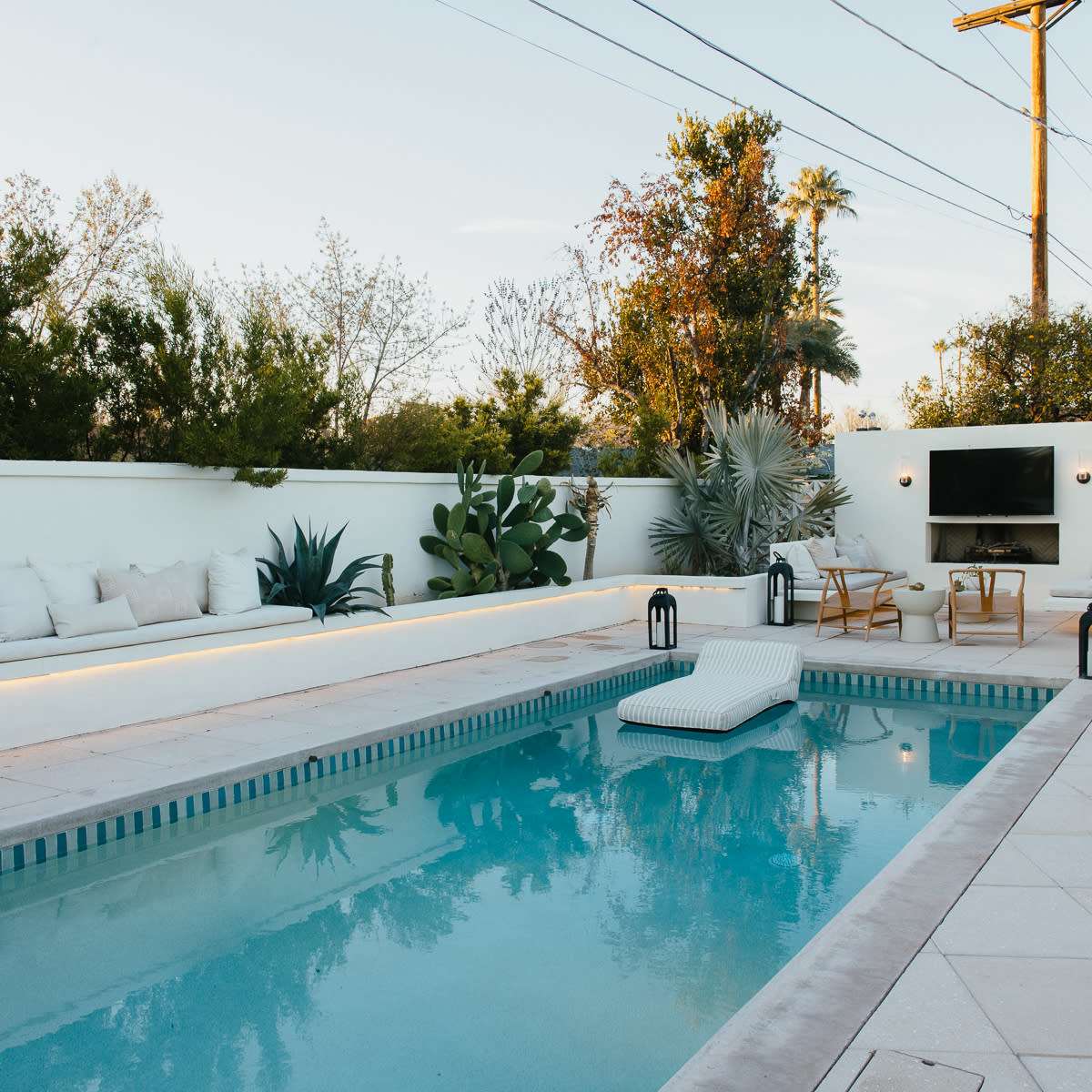 A modern outdoor pool area with a lounge seating space, potted plants, and a television mounted on a wall.