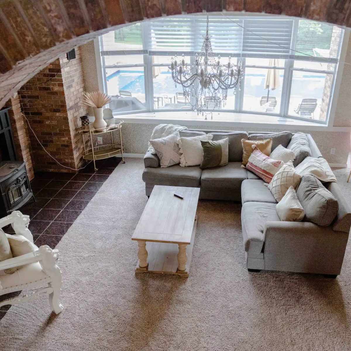 A spacious living room features a large sectional sofa, a coffee table, and a chandelier, with large windows overlooking a pool area.
