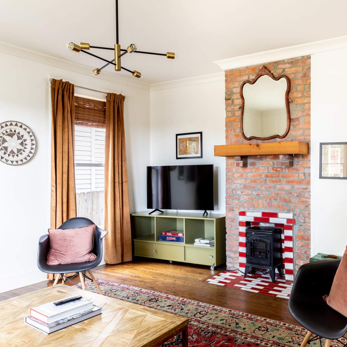 A modern living room featuring a brick fireplace, a television, two black chairs, a wooden coffee table, and large windows with curtains.