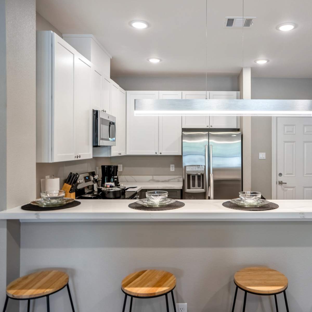 A modern kitchen features a counter with four wooden stools, sleek white cabinetry, and stainless steel appliances.