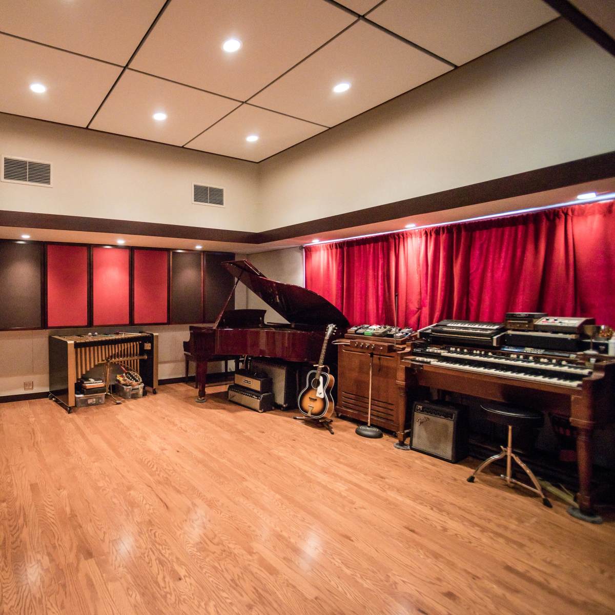 A music studio featuring a grand piano, various keyboards, a vibraphone, an electric guitar, and several vintage audio equipment arranged in a well-lit room with red curtains.