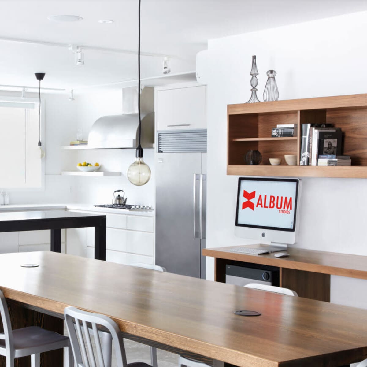 A modern kitchen with a large wooden dining table, sleek appliances, and a computer on a desk in the corner.