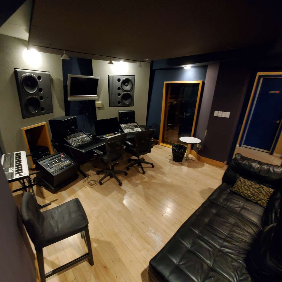 A recording studio featuring two mixing stations, large speakers mounted on the walls, a keyboard, a couch, and a small table.
