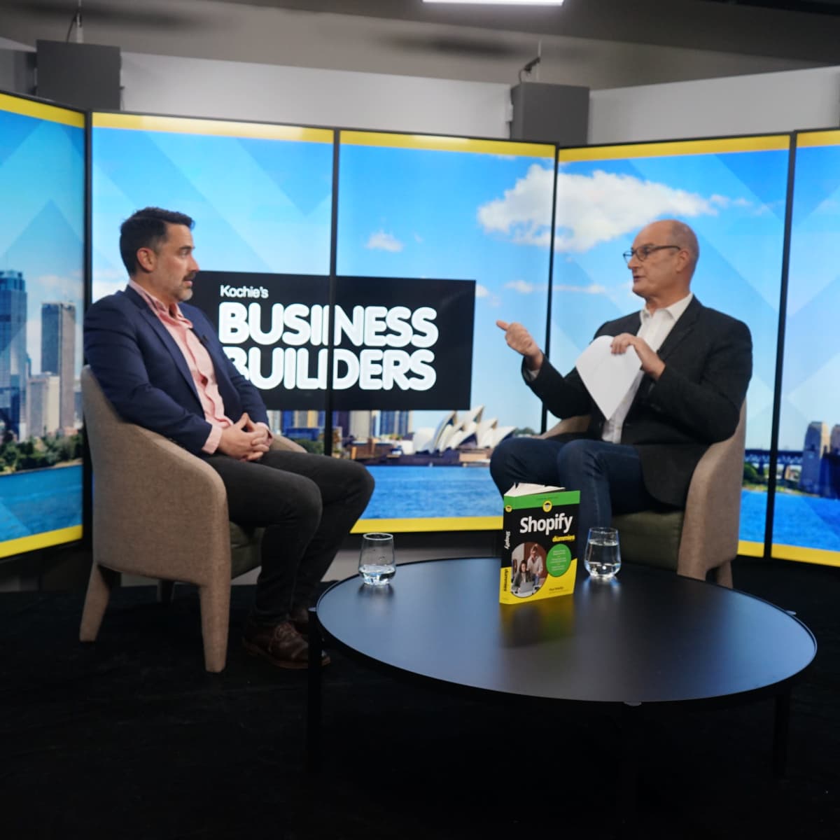 A television studio setting features two men seated in chairs, discussing in front of a backdrop of a city skyline, with a promotional book for Shopify on the table.