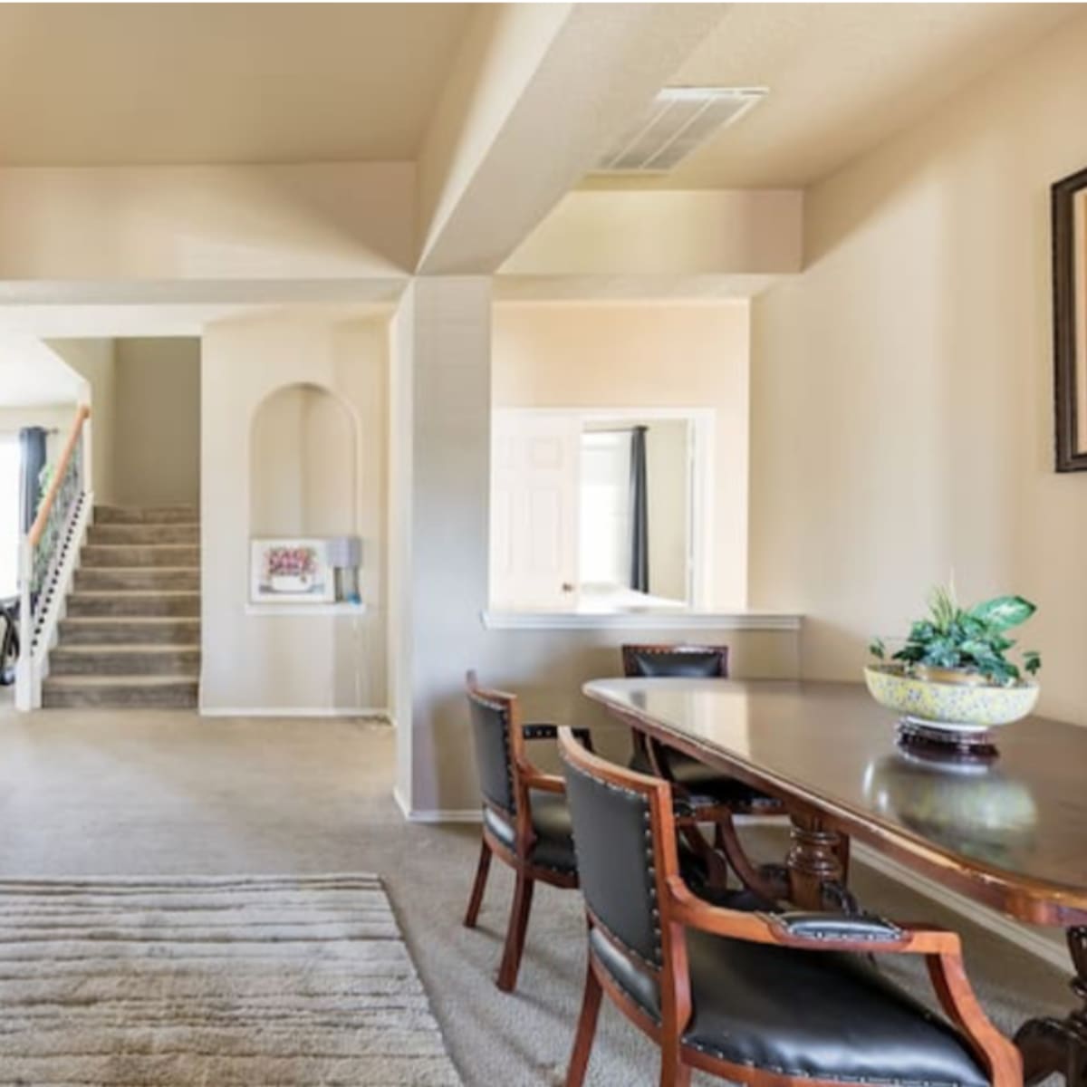 A hallway with a staircase, a dining table with chairs, a large mirror leaning against the wall, and light-colored walls.