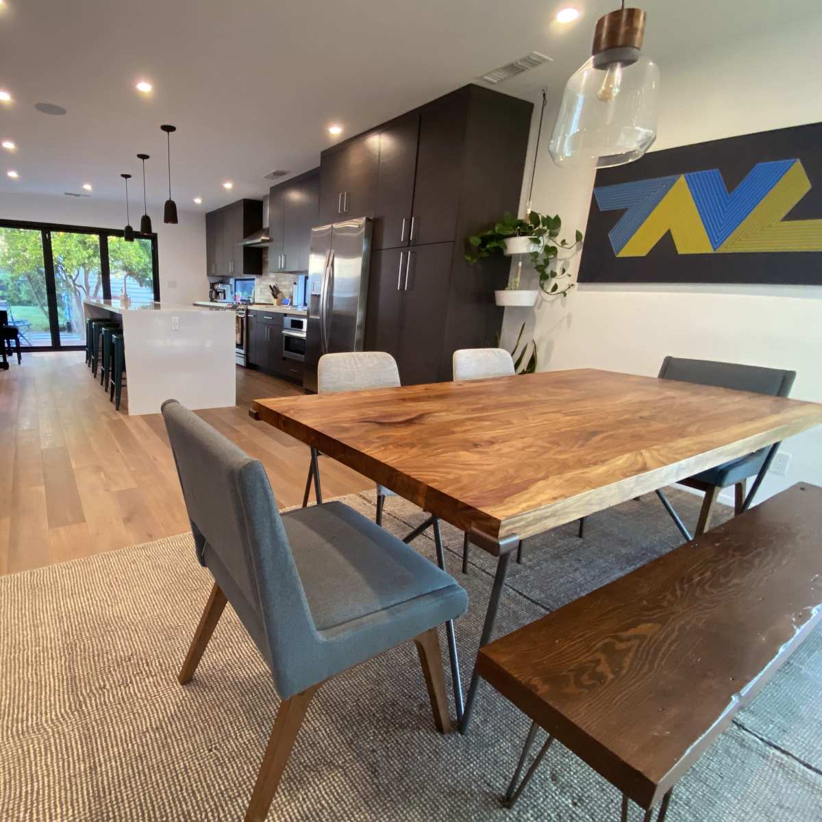 A modern open-concept living and dining area featuring a wooden dining table, gray chairs, and a kitchen in the background with large glass doors leading to an outdoor space.