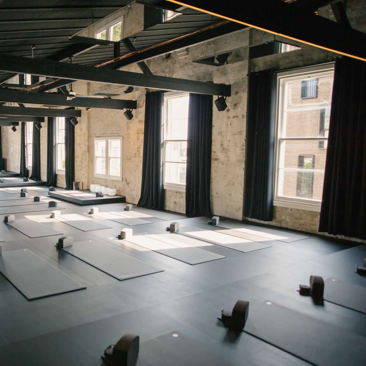A spacious yoga studio features multiple exercise mats arranged on a dark floor, with large windows allowing natural light to enter.