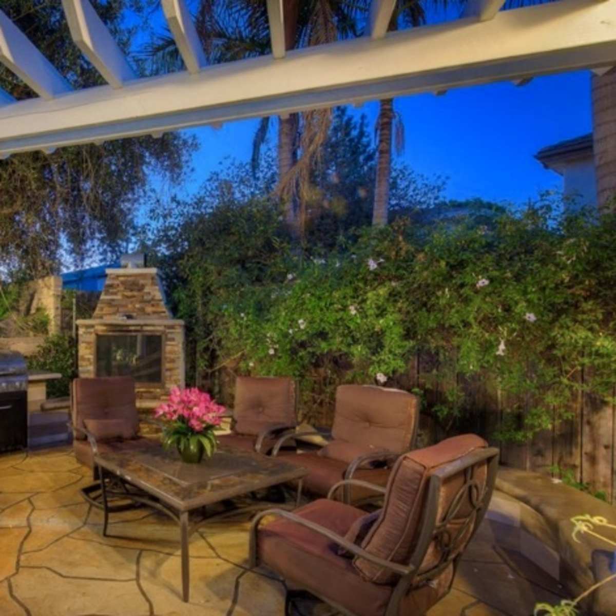 A shaded patio features a stone table surrounded by four chairs, with a barbecue grill and stone fireplace visible nearby.