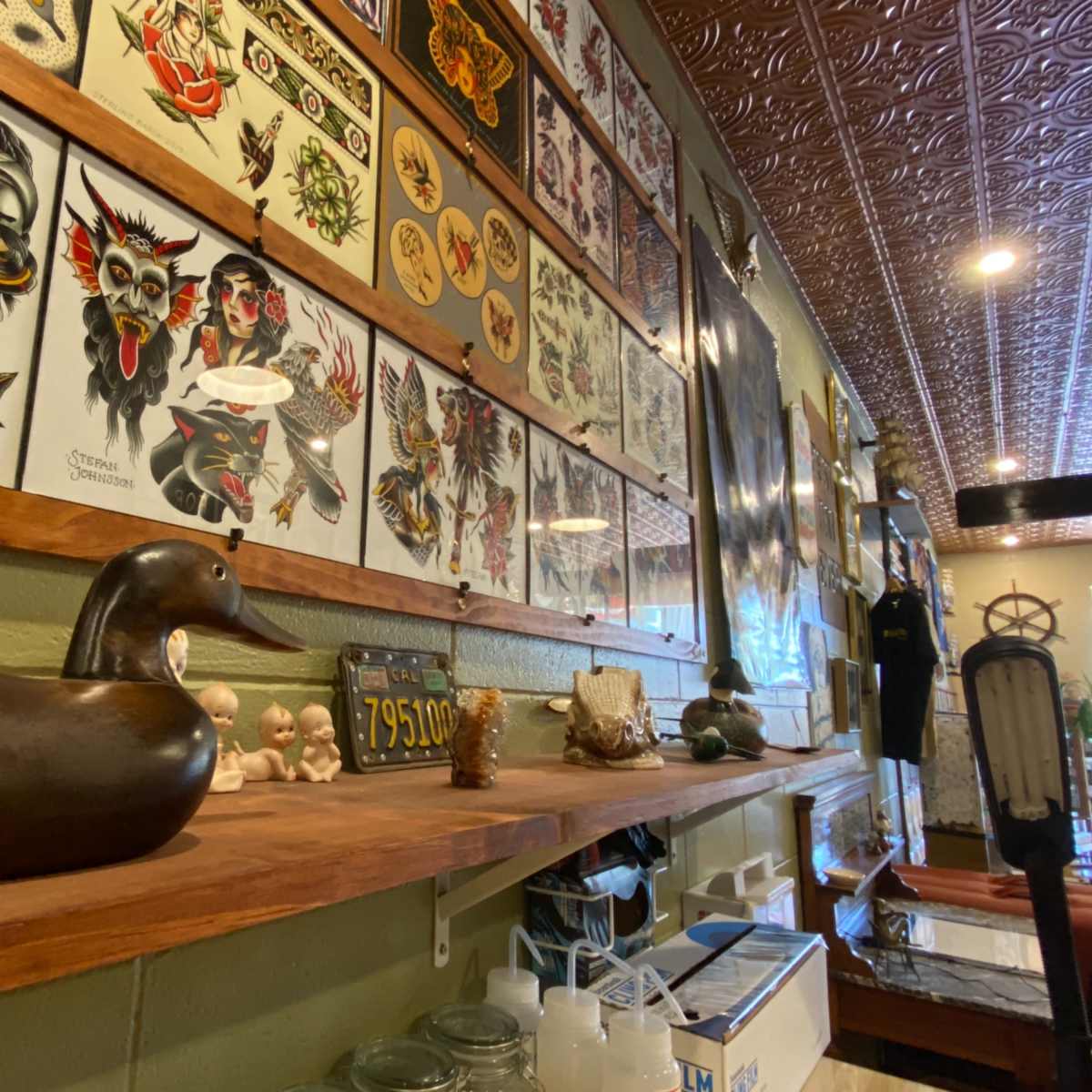A wooden shelf displaying various decorative items, including a carved duck, small figurines, and glass containers, alongside a wall adorned with tattoo flash art.