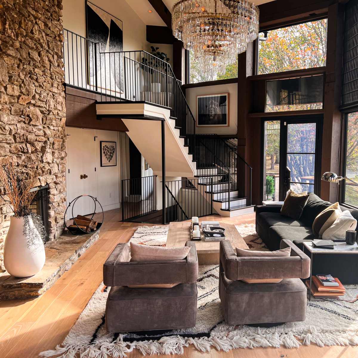 A modern living room with a stone fireplace, large windows, a staircase, and contemporary furniture arranged around a central rug.