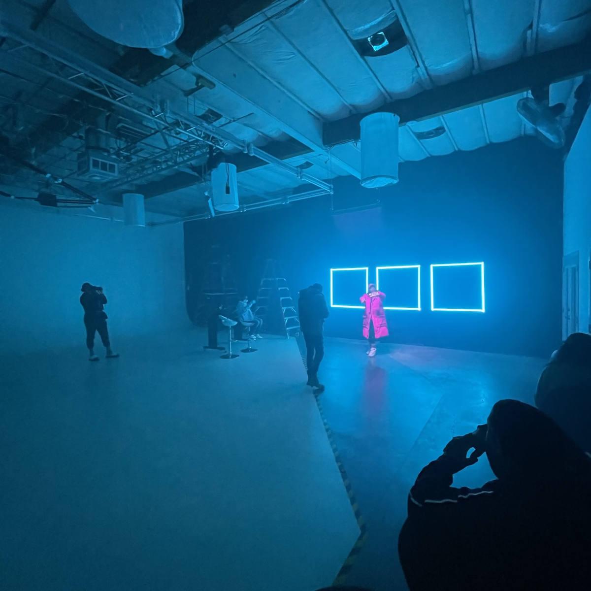 A person in a bright outfit poses in front of illuminated rectangular panels while several others observe and photograph the scene in a spacious studio.
