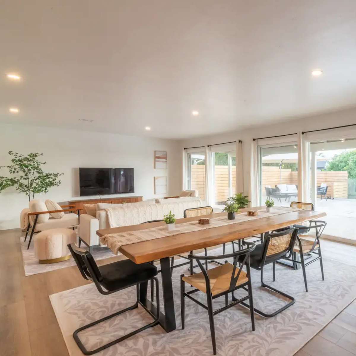 A spacious living and dining area features a wooden table surrounded by black chairs, an exposed brick wall, and large windows leading to an outdoor space.