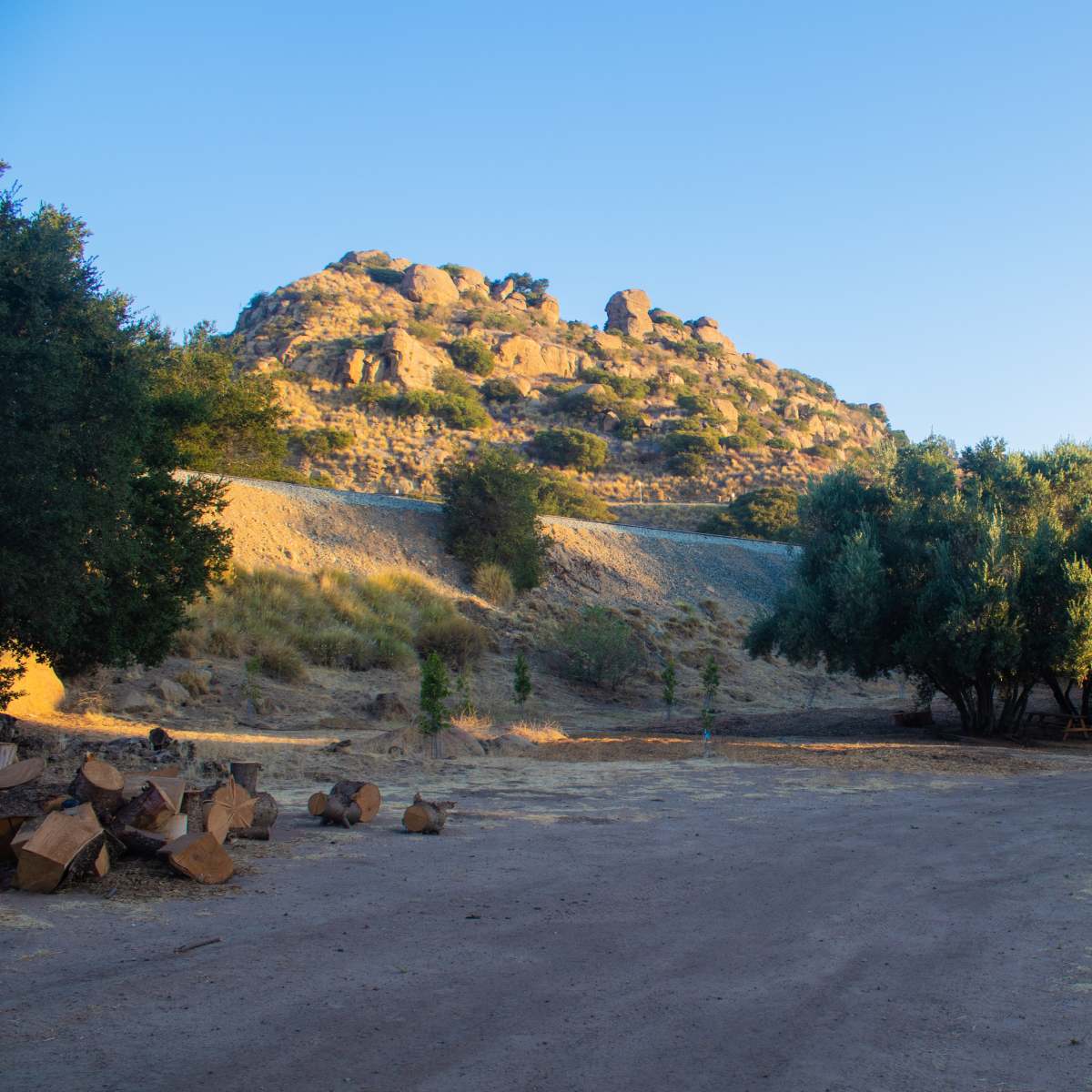 A dirt path leads through a sparse landscape with a rocky hill and clusters of trees under a clear sky.