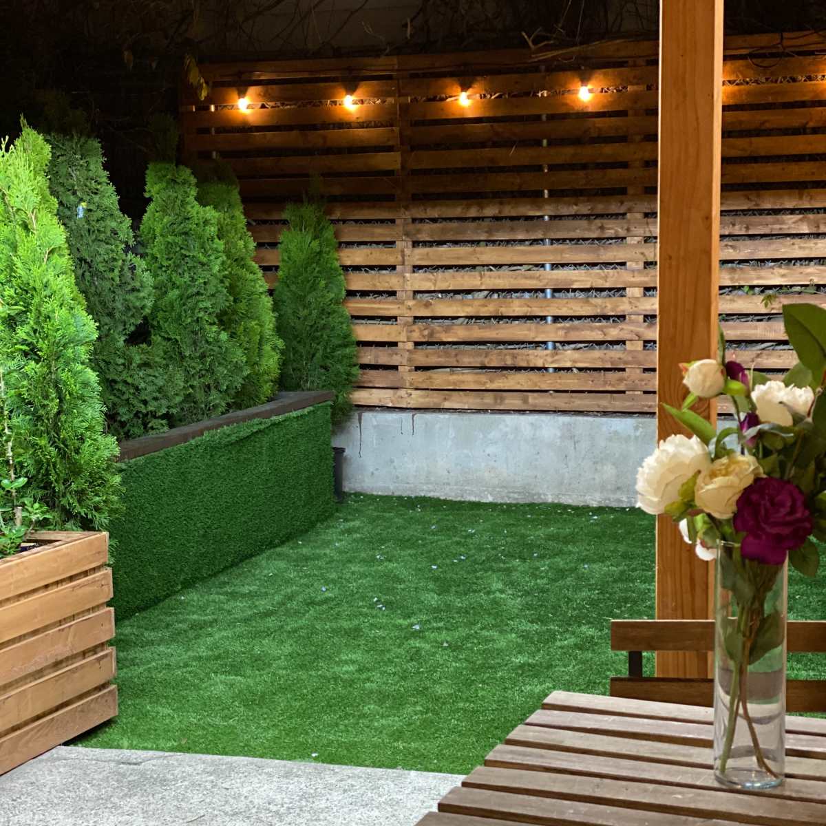 A small outdoor space features artificial grass, wooden fencing, and potted plants, with a table set on a concrete surface.
