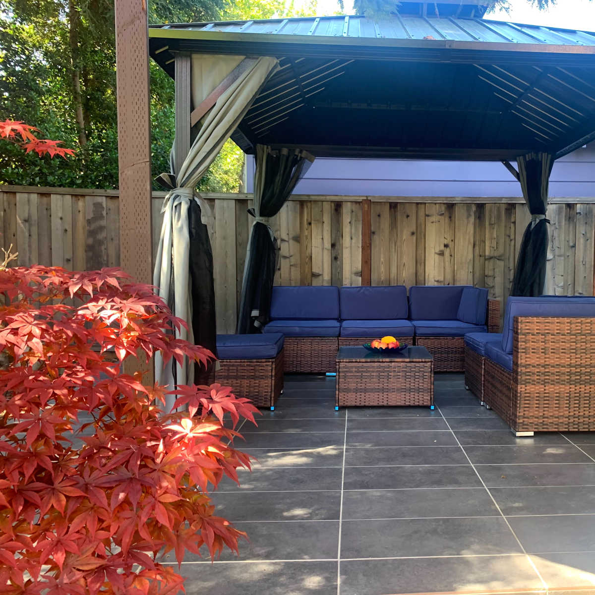 A gazebo contains a seating area with wicker furniture and a small table, surrounded by greenery and a wooden fence.
