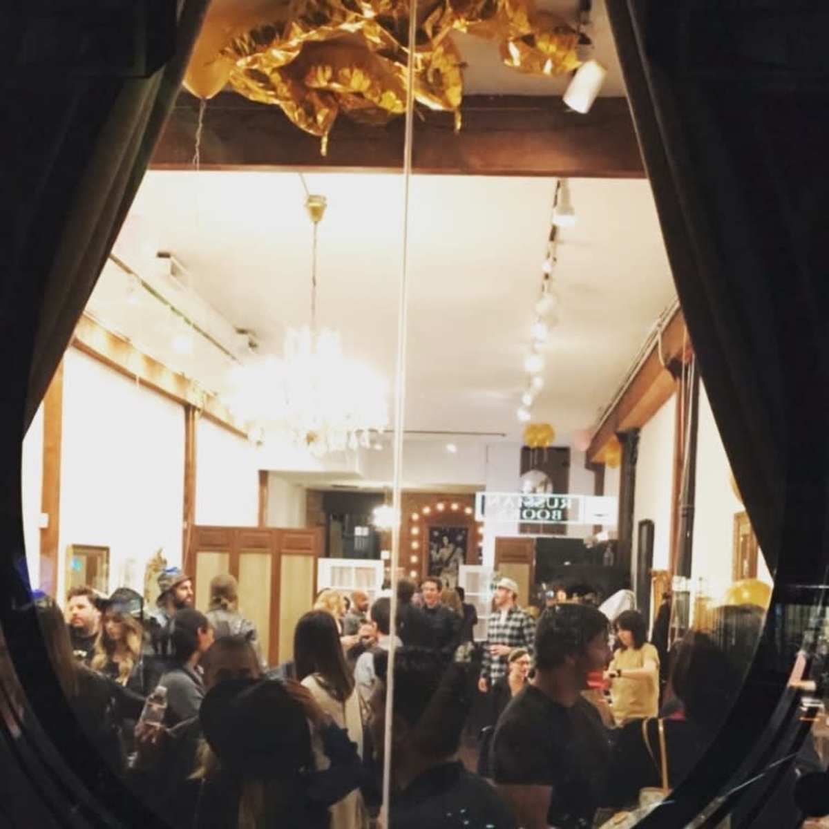 A crowded indoor gathering with people mingling under decorative lights and a chandelier.