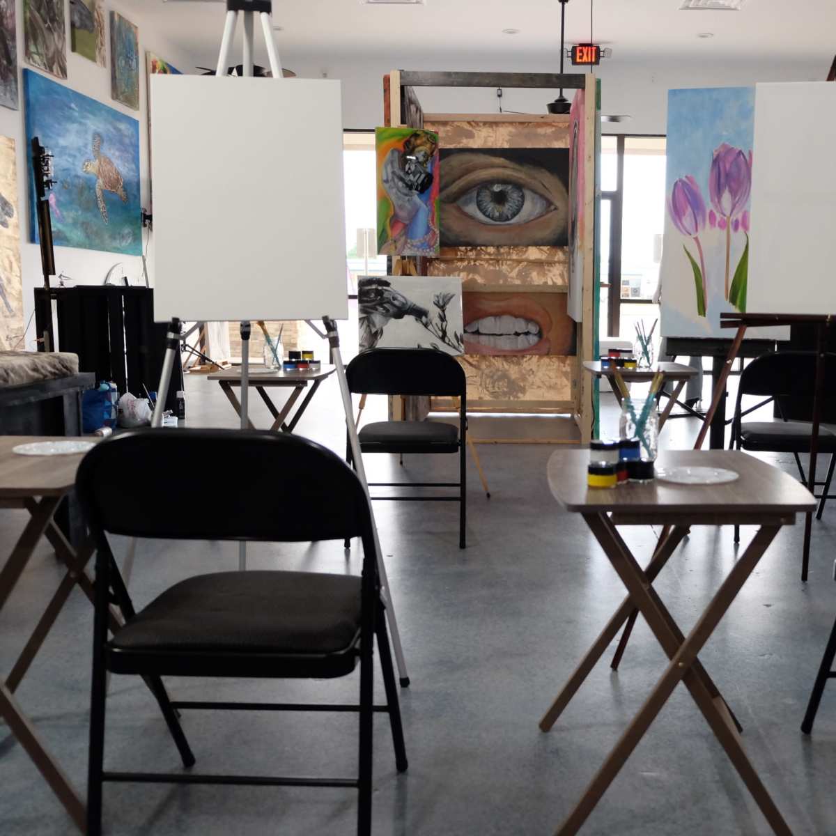 An art studio with empty tables and chairs arranged in front of blank canvases, surrounded by colorful artwork on the walls.